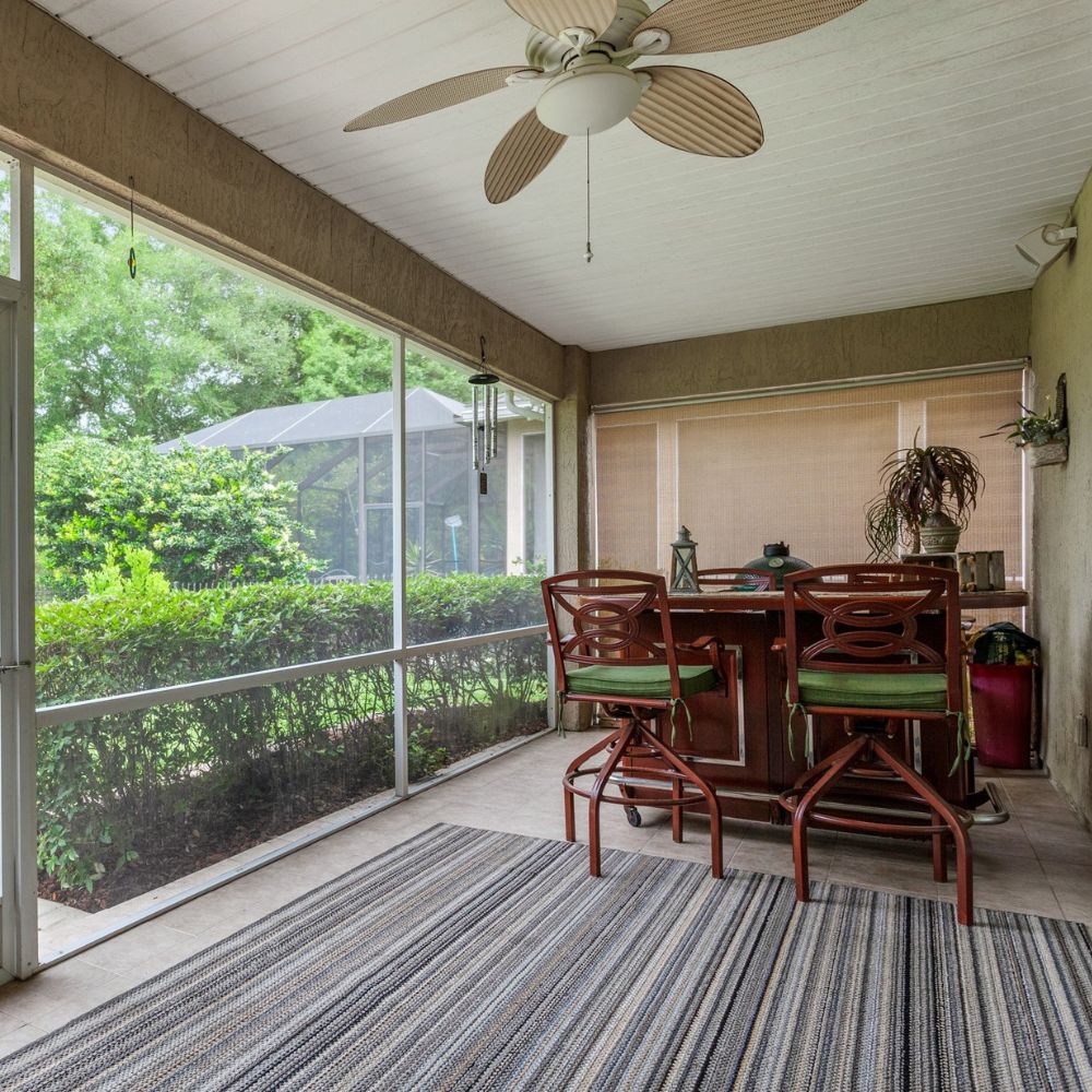 The interior of a screened-in porch with a striped rug and bar with two barstools inside on the right and a yard with bushes and trees outside of the screen wall and door on the left.
